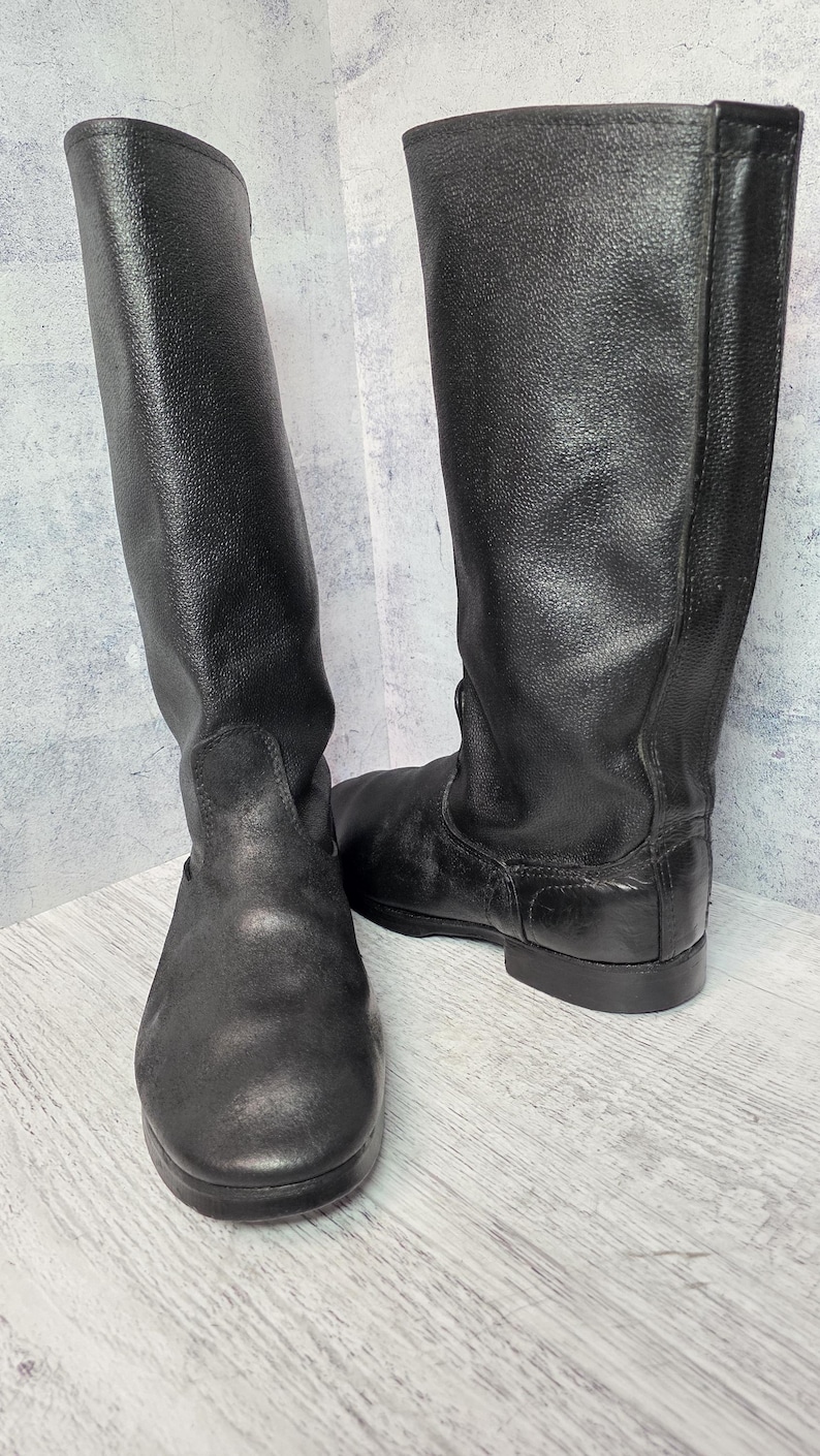 Puede incluir: Un par de botas negras de cuero hasta la rodilla. Las botas tienen un acabado liso y mate y una puntera redondeada. Las suelas son planas y las botas parecen sin usar. Las botas est&aacute;n de pie sobre un fondo gris claro.