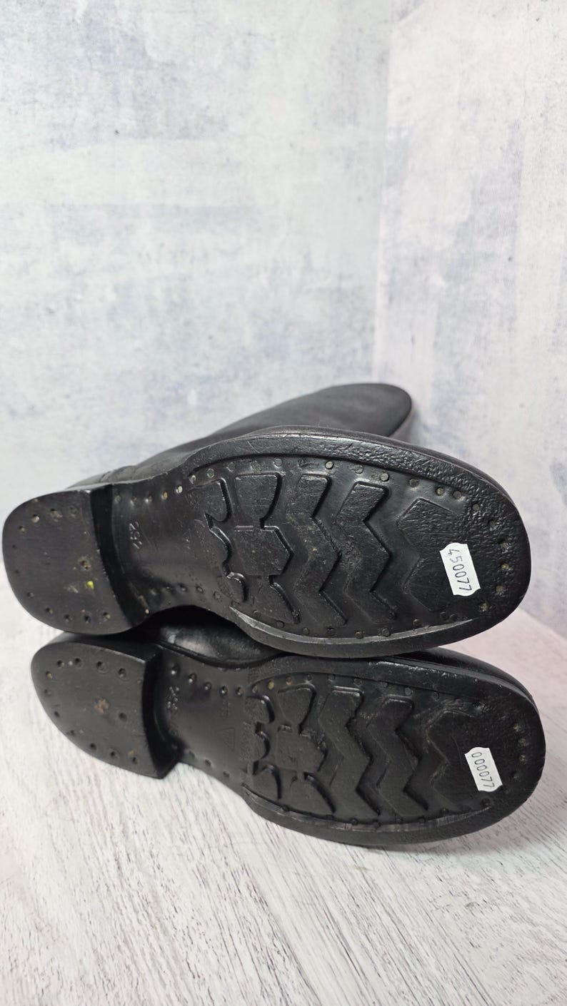 Puede incluir: Botas de cuero negro con suela texturizada. Las suelas tienen un patr&oacute;n distintivo para el agarre y est&aacute;n marcadas con una pegatina blanca. Las botas son probablemente para uso en exteriores y son duraderas.