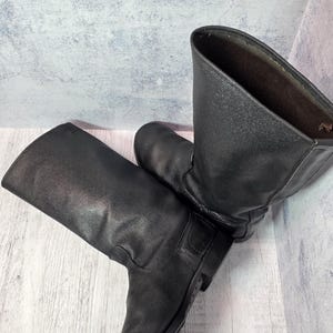 Puede incluir: Un par de botas de cuero negro con ca&ntilde;a hasta la rodilla. Las botas tienen un dise&ntilde;o cl&aacute;sico con punta redonda y tac&oacute;n bajo. El cuero parece ligeramente desgastado, d&aacute;ndoles un aspecto vintage.