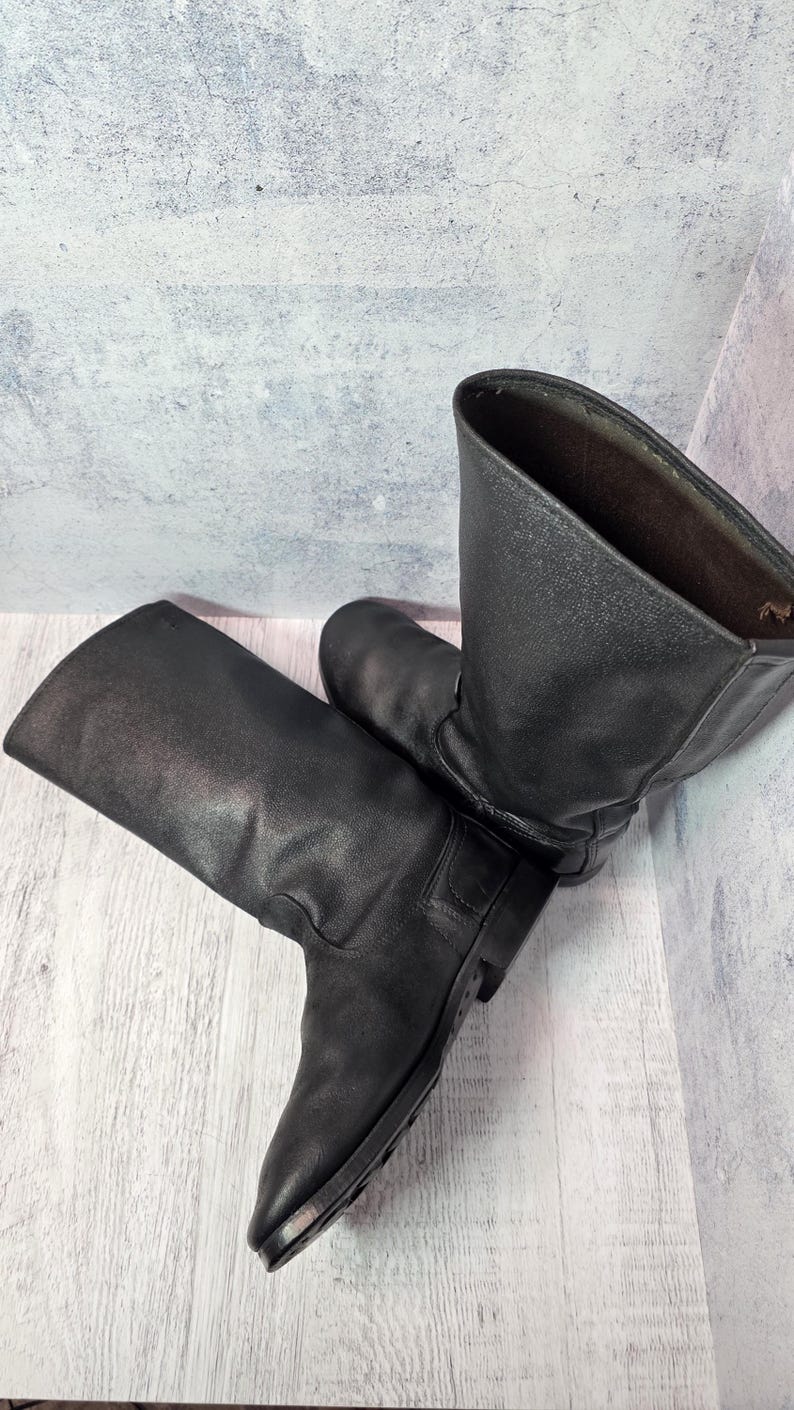 Puede incluir: Un par de botas de cuero negro con ca&ntilde;a hasta la rodilla. Las botas tienen un dise&ntilde;o cl&aacute;sico con punta redonda y tac&oacute;n bajo. El cuero parece ligeramente desgastado, d&aacute;ndoles un aspecto vintage.