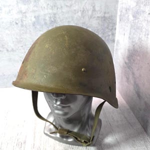 May include: An olive drab military helmet with a chin strap. The helmet is displayed on a silver mannequin head. The helmet has a slightly weathered appearance, suggesting age or use. The background is a light gray and white.