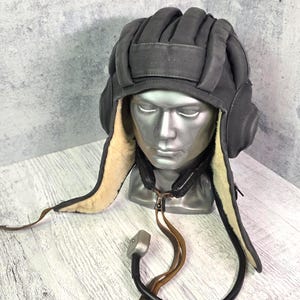 Könnte beinhalten: Grauer Panzerhelm mit Ohrenklappen und gepolstertem Oberteil, ausgestellt auf einem Schaufensterkopf. Das Innenfutter ist hellbraun. Ein schwarzes und braunes Kommunikationsgerät ist am Helm befestigt.