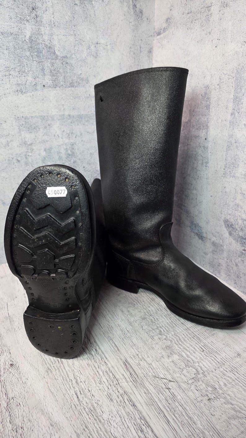 Puede incluir: Un par de botas de cuero negro. Una bota est&aacute; de pie, mostrando la ca&ntilde;a alta, mientras que la otra est&aacute; tumbada de lado, revelando la suela con un patr&oacute;n texturizado y una etiqueta con el n&uacute;mero "450077".