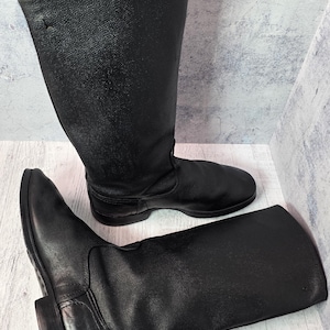 Puede incluir: Un par de botas de cuero negro. Las botas son altas, llegando a la mitad de la pantorrilla, con punta redonda y tac&oacute;n bajo. El cuero tiene un aspecto texturizado. Una bota est&aacute; de pie, la otra est&aacute; tumbada de lado.
