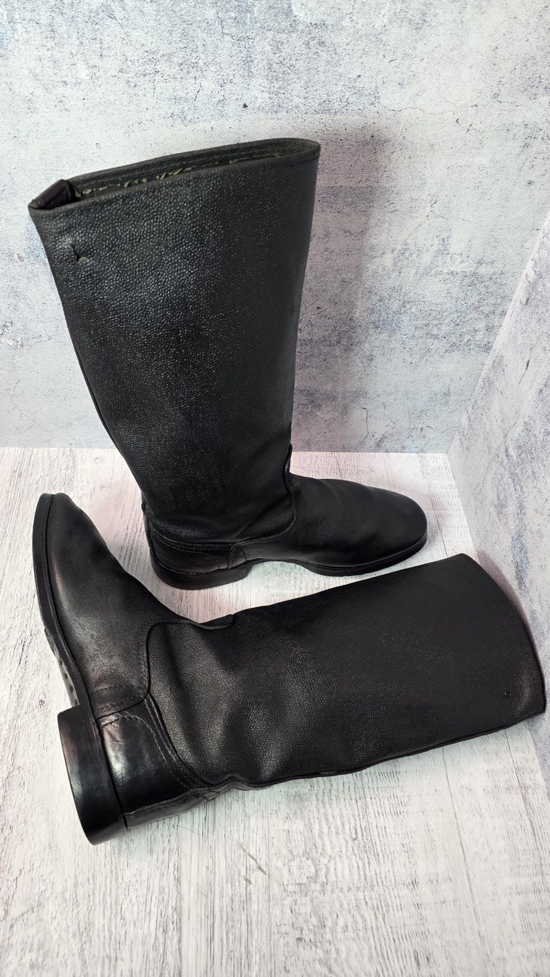 Puede incluir: Un par de botas de cuero negro. Las botas son altas, llegando a la mitad de la pantorrilla, con punta redonda y tac&oacute;n bajo. El cuero tiene un aspecto texturizado. Una bota est&aacute; de pie, la otra est&aacute; tumbada de lado.