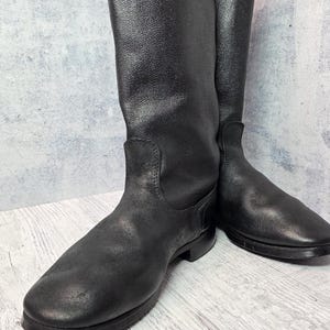 Puede incluir: Un par de botas negras de cuero hasta la rodilla. Las botas tienen un acabado liso y mate y una puntera redondeada. La ca&ntilde;a de las botas es alta y recta, y las suelas son planas. Las botas se exhiben sobre un fondo neutro.