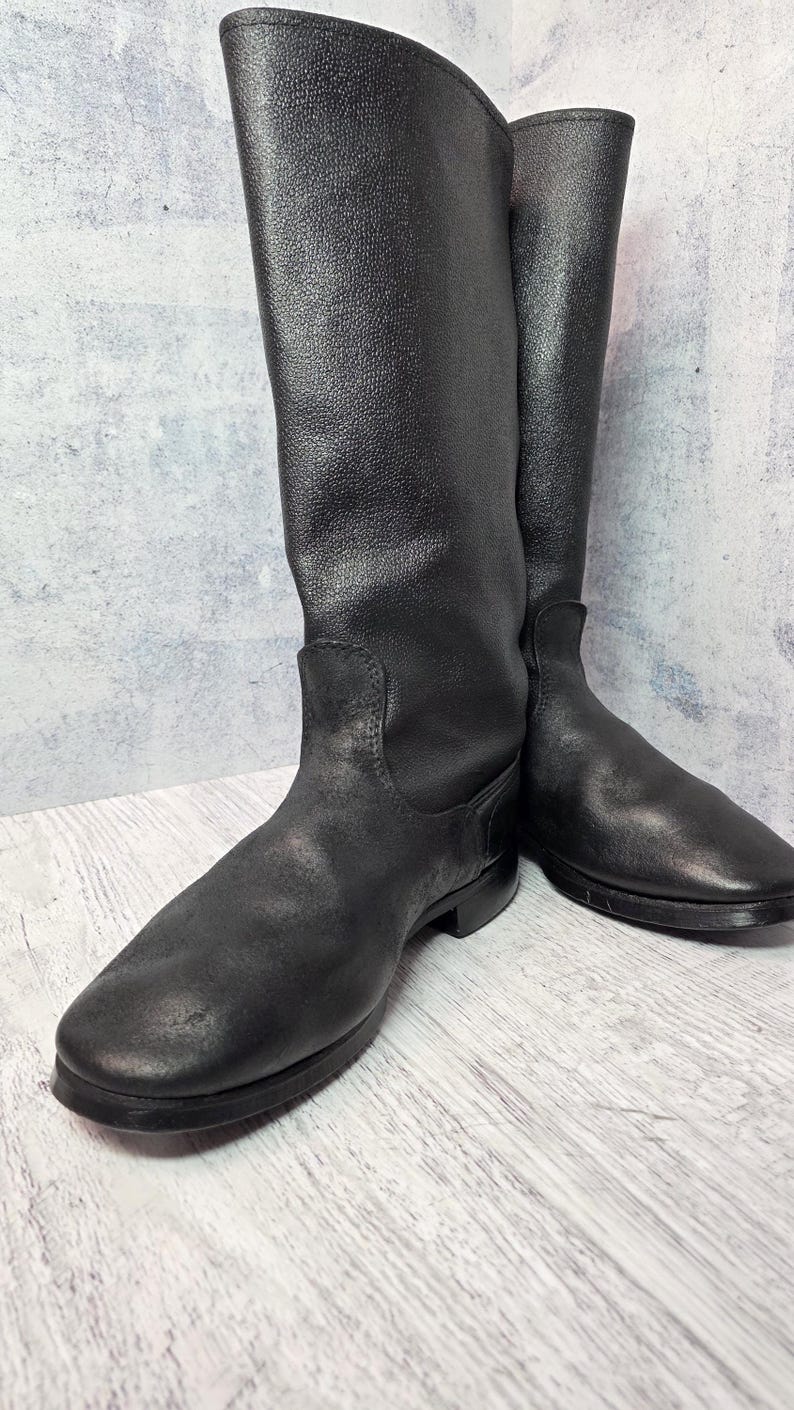 Puede incluir: Un par de botas negras de cuero hasta la rodilla. Las botas tienen un acabado liso y mate y una puntera redondeada. La ca&ntilde;a de las botas es alta y recta, y las suelas son planas. Las botas se exhiben sobre un fondo neutro.