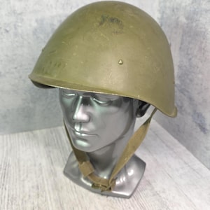 Può includere: Un elmetto militare color verde oliva caratterizzato da una forma a cupola liscia e arrotondata con una tesa corta. L'elmetto include un sottogola in tessuto color marrone chiaro con fibbia in metallo ed è esposto su un manichino argentato.