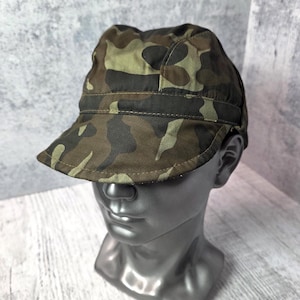 May include: Camouflage pattern cap in shades of green, brown, and black. The cap features a short brim and a structured crown. The cap is displayed on a mannequin head, showcasing its shape and design.
