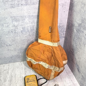 Puede incluir: Un contenedor de tela naranja tipo paracaídas con correas blancas que llevan el texto "MAP-2M No 0888569". Al lado hay un dispositivo electrónico rectangular amarillo etiquetado como "Прибой-2с" con un cable negro y un imperdible metálico.