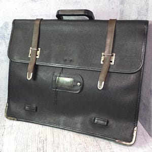 Könnte beinhalten: Eine schwarze Aktentasche mit strukturierter Oberfläche, ausgestattet mit zwei braunen vertikalen Riemen und silberfarbenen Metallschnallen. Die Tasche verfügt über einen schwarzen Tragegriff, ein zentrales rechteckiges Namensschild und silberne Metallecken.