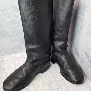Puede incluir: Un par de botas altas de cuero negro. Las botas tienen un acabado liso y mate y un dise&ntilde;o cl&aacute;sico, con una puntera redondeada y una ca&ntilde;a alta que llega hasta la mitad de la pantorrilla. Las botas est&aacute;n sobre un fondo neutro.
