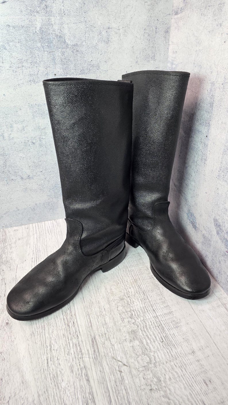Puede incluir: Un par de botas altas de cuero negro. Las botas tienen un acabado liso y mate y un dise&ntilde;o cl&aacute;sico, con una puntera redondeada y una ca&ntilde;a alta que llega hasta la mitad de la pantorrilla. Las botas est&aacute;n sobre un fondo neutro.