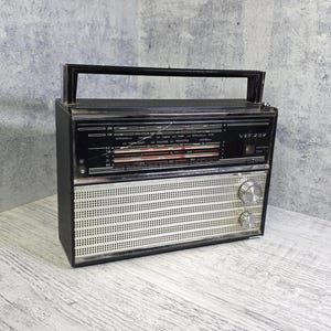 Puede incluir: Una radio portátil negra con una rejilla de altavoz de metal plateado y un asa de transporte superior. El panel frontal presenta una pantalla negra con texto en blanco y rojo, incluyendo el modelo VEF 202, y dos perillas de control plateadas.