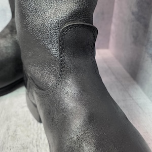 Puede incluir: Primer plano de un par de botas de cuero negro. Las botas tienen una superficie texturizada y una puntera redondeada. La ca&ntilde;a de la bota se extiende hacia arriba y las costuras son visibles. Las botas son probablemente para uso en exteriores.