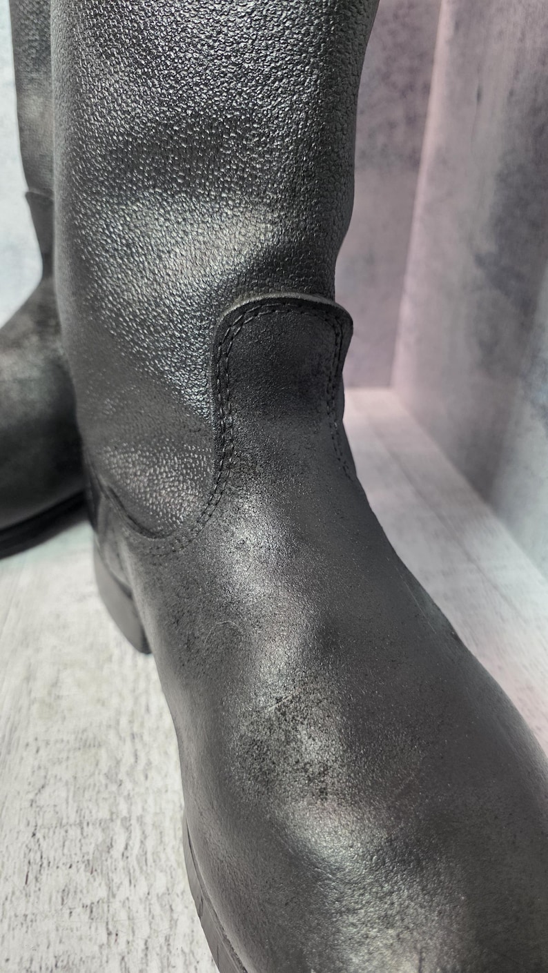 Puede incluir: Primer plano de un par de botas de cuero negro. Las botas tienen una superficie texturizada y una puntera redondeada. La ca&ntilde;a de la bota se extiende hacia arriba y las costuras son visibles. Las botas son probablemente para uso en exteriores.