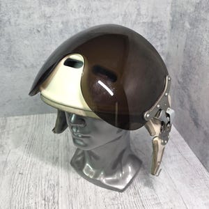 Könnte beinhalten: Ein futuristisch anmutender Helm mit dunkelbraunem Visier und cremefarbenem Sockel. Der Helm verfügt über einen komplexen metallischen Scharniermechanismus an der Seite. Der Helm wird auf einem silbernen Schaufensterkopf präsentiert.
