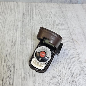 Può includere: Un esposimetro vintage in una custodia di pelle marrone. L'esposimetro ha un quadrante nero con segni bianchi e rossi e un quadrante color argento. La custodia è aperta, rivelando il quadrante dell'esposimetro. Lo sfondo è una superficie di legno di colore chiaro.