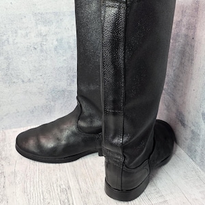 Puede incluir: Un par de botas negras de cuero hasta la rodilla. Las botas tienen un acabado liso y mate y un dise&ntilde;o cl&aacute;sico. Las suelas son planas y las botas parecen sin usar. Las botas miden aproximadamente 40 cm de altura.