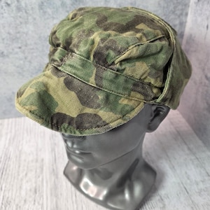 Op de afbeelding: Een camouflagepatroon pet in groen- en bruintinten, weergegeven op een mannequinhoofd. De pet heeft een korte klep en een ronde kroon. De stof lijkt duurzaam katoen of iets dergelijks te zijn. De pet is een accessoire in militaire stijl.