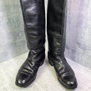 Puede incluir: Un par de botas altas de cuero negro hasta la rodilla con acabado liso, puntera redondeada y tacón bajo. Las botas presentan una caña alta y estructurada que llega a la rodilla, con costuras visibles alrededor del pie y un diseño sencillo.