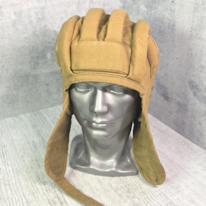 May include: A tan-coloured, padded protective helmet featuring a series of vertical, cushioned ridges across the crown and extended side flaps that hang down. The helmet is constructed from a durable fabric material and includes a chin strap.