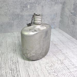 May include: A silver-coloured canteen with a screw-on cap and a chain. The canteen has a rounded rectangular shape and is made of metal. The cap is attached to the canteen with a small chain. The canteen is on a white wooden surface.