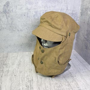 Op de afbeelding: Een beige canvas pet met een brede klep en een geïntegreerde gezichtsbedekking die naar beneden loopt om de nek en kin te bedekken. Het mondkapje is bevestigd met kleine, ronde, donkergekleurde knopen en is voorzien van zijflappen.