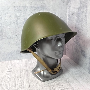 Könnte beinhalten: Ein olivgrüner Militärhelm mit abgerundeter Krone und Krempe. Der Helm wird auf einem grauen Schaufensterkopf mit einem beigefarbenen Kinnriemen präsentiert. Der Hintergrund ist eine strukturierte graue Wand und eine weiße Holzoberfläche.