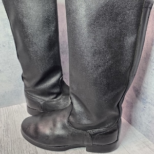 Puede incluir: Un par de botas negras de cuero hasta la rodilla. Las botas tienen un acabado liso y mate y una puntera redondeada. Las suelas son planas y las botas parecen sin usar. Las botas est&aacute;n de pie sobre un fondo gris claro.