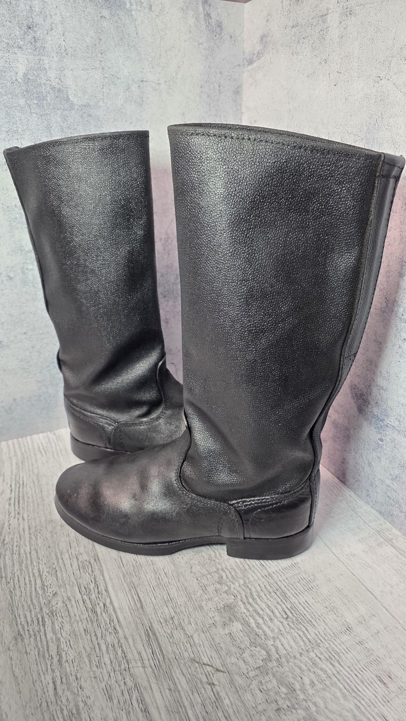 Puede incluir: Un par de botas negras de cuero hasta la rodilla. Las botas tienen un acabado liso y mate y una puntera redondeada. Las suelas son planas y las botas parecen sin usar. Las botas est&aacute;n de pie sobre un fondo gris claro.
