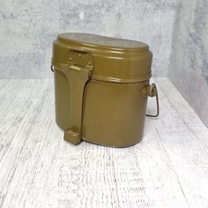 May include: Olive green metal military-style mess kit with a hinged lid and a folding handle. The kit has a cylindrical body and a rectangular handle. The kit is designed for outdoor use and camping.