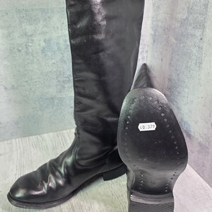 Puede incluir: Un par de botas altas de cuero negro con acabado liso y punta redondeada. Una bota está de pie, mientras que la otra está colocada para mostrar la suela negra, que tiene una pegatina blanca con el texto impreso 40.379.