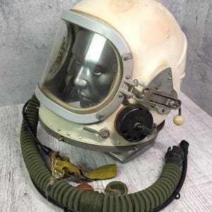 Può includere: Casco spaziale vintage color crema con visiera trasparente, dettagli in metallo e un tubo scanalato verde. Il casco ha una superficie riflettente all'interno della visiera e un quadrante nero con il numero 2031.
