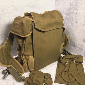 May include: Olive green canvas backpack with multiple pockets and adjustable straps. Includes two smaller matching pouches. The bag has a flap closure and appears to be vintage or military-style.