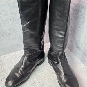 May include: A pair of tall, black leather boots. The boots have a smooth, shiny finish and a classic design. They are knee-high with a rounded toe and a low heel. The boots are standing on a light-coloured wooden surface.