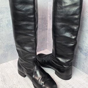 May include: A pair of black leather knee-high boots. The boots have a smooth, polished finish and a flat sole. The shaft of the boots is tall and straight, with a slight curve at the top. The boots are displayed on a light-colored wooden surface.