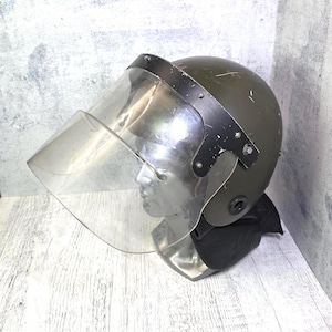 Può includere: Un casco antisommossa verde oliva dotato di una grande visiera curva e trasparente in policarbonato fissata con una montatura in metallo nero. Il casco include una protezione per il collo nera alla base ed è esposto su una testa di manichino argentata.