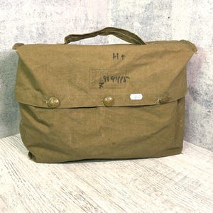 Op de afbeelding: Een olijfgroene canvas tas met een handvat aan de bovenzijde en een overslagklep die wordt vastgezet met drie ronde metalen knopen. De klep bevat zwarte inktmarkeringen, waaronder de tekst BKK-6M en 84144/5, en een klein wit rechthoekig label.