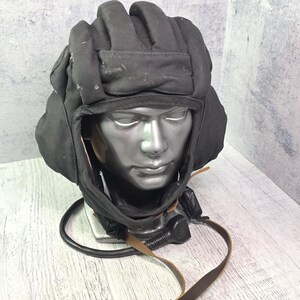 May include: A black padded tank helmet featuring a series of horizontal protective rolls across the top, integrated ear flaps, and a chin strap with a brown leather adjustment buckle. A black communication cable and connector extend from the base.
