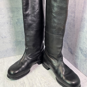 Puede incluir: Un par de botas altas de cuero negro con punta redondeada, caña lisa y tacón bajo de bloque. Las botas presentan detalles de costuras visibles alrededor del talón y el tobillo, con un acabado de grano texturizado en toda la superficie.