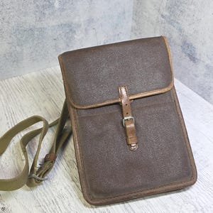 Könnte beinhalten: Braune Leder-Umhängetasche mit Klappenverschluss und Schnalle. Die Tasche hat eine strukturierte Oberfläche und eine kontrastierende braune Umrandung. Ein olivgrüner Canvas-Riemen ist befestigt.