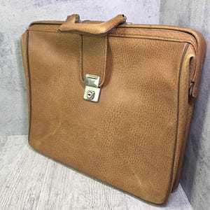 May include: A tan leather briefcase with a textured surface, featuring a single top handle and a central silver-toned metal locking mechanism. The rectangular bag has rounded edges and a structured frame, showcasing a classic design with visible stitching.
