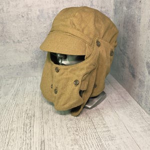 May include: A tan-colored cap with a stiff brim and a detachable face mask covering the lower portion. The cap features small circular buttons along the sides and front, securing the mask to the main body of the headwear. The fabric has a textured appearance.