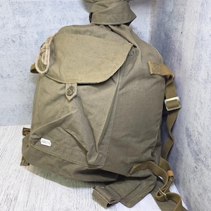 Puede incluir: Una mochila de lona verde oliva con cierre de cordón, un bolsillo frontal con solapa y botón, y correas ajustables de cincha. Una pequeña etiqueta rectangular blanca con el número impreso 000350 está adherida en la parte frontal.