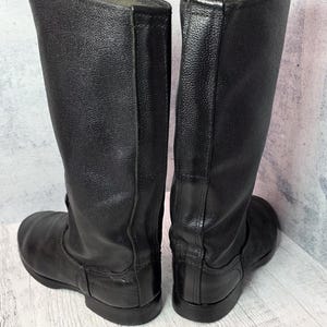 Puede incluir: Un par de botas altas de cuero negro. Las botas tienen una punta redondeada, una suela plana y una ca&ntilde;a alta. El cuero parece ser texturizado. Las botas se exhiben sobre un fondo neutro.