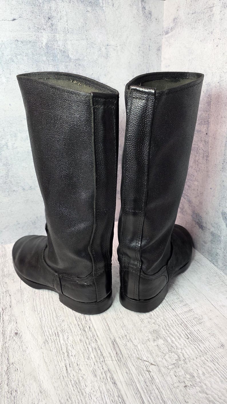 Puede incluir: Un par de botas altas de cuero negro. Las botas tienen una punta redondeada, una suela plana y una ca&ntilde;a alta. El cuero parece ser texturizado. Las botas se exhiben sobre un fondo neutro.
