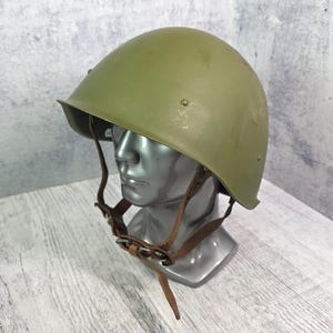 May include: An olive green military-style helmet with a smooth, rounded dome and a short front brim. The helmet features a brown leather chin strap with metal buckles and rivets, secured around a silver-toned, featureless mannequin head display.
