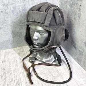 May include: A grey tank helmet with ear flaps and a chin strap, displayed on a mannequin head. The helmet has a padded design and a leather strap with a buckle. A black cable extends from the helmet.