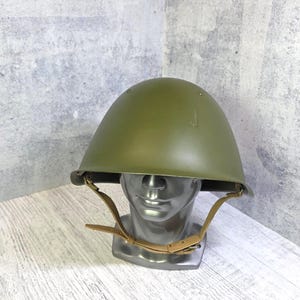 May include: An olive green military helmet with a leather chin strap. The helmet is displayed on a silver mannequin head against a textured grey and white background. The helmet is a classic military style.
